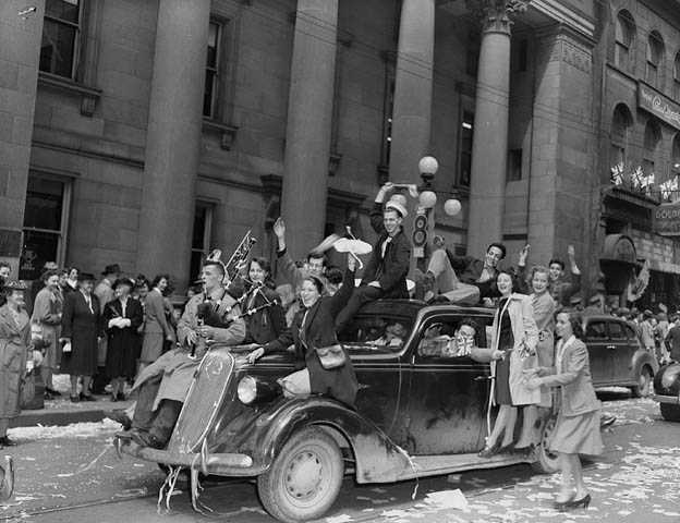 Black and white photograph. A car drives down the street over tickertape. Happy people ride inside and on top of it. Others crowd the street in front of a large concrete building.